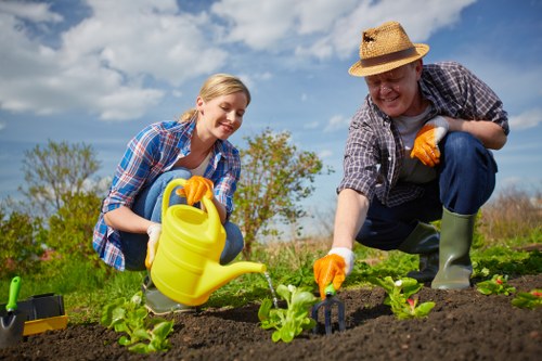 A woman and an older man are working together in a well-maintained garden in Sanderstead, planting young green shoots in dark fertile soil bordered by neat garden beds. The woman is wearing a checked shirt, smiling as she holds a yellow watering can, ready to water the newly planted plants. The man, wearing a straw hat, a plaid shirt, and waterproof boots, is kneeling on the garden bed, carefully tending to the plants. The garden features a spacious lawn with lush, green grass in the background, alongside a hedge line and some mature trees under a partly cloudy sky. The scene captures outdoor gardening activity with natural daylight illuminating the area, highlighting the orderly planting beds, the rich soil, and the vibrant green foliage. This setting reflects the kind of outdoor space that professional gardening services in Sanderstead might help maintain, including planting, lawn care, and general garden upkeep, offering a peaceful residential garden environment in early spring or summer.